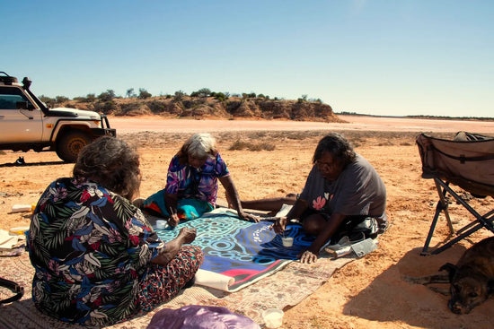 Three women sitting on the ground outdoors with a vehicle and landscape in the background