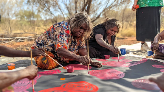 Indigenous artists creating a design on a large canvas outdoors.